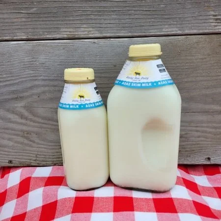 5e6e71e4-1ece-4e41-a797-4df5430dba19 Two bottles of fresh milk, one large and one small, with labels indicating they contain whole milk, placed on a red and white checkered tablecloth against a wooden backdrop.