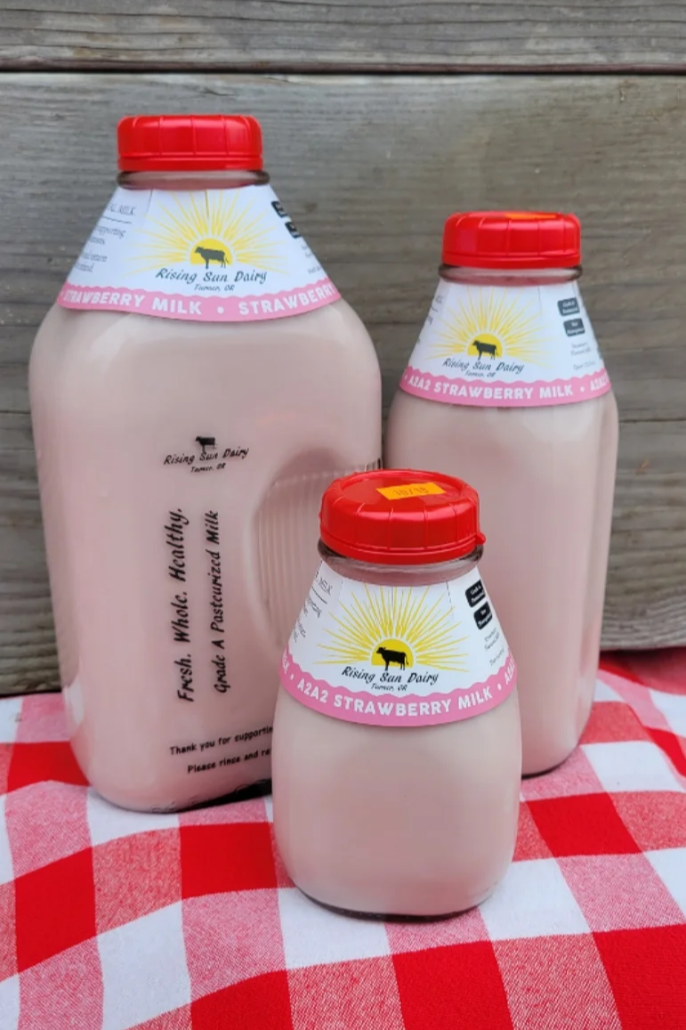 strawberry+clip Three bottles of Rising Sun Dairy strawberry milk in varying sizes, featuring colorful labels that highlight their fresh and healthy qualities. The bottles are displayed on a red and white checkered tablecloth, showcasing the brand's commitment to quality dairy products.